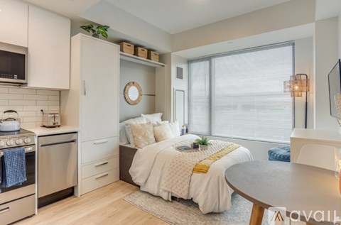 A bedroom with a bed, a table, and a kitchen area with a microwave and oven.