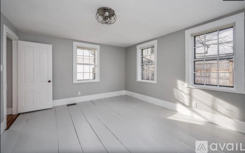 A room with white flooring and grey walls has a white door and two windows.