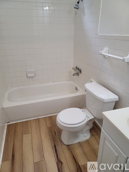 A white toilet sits in a bathroom with a white tub and wooden floors.