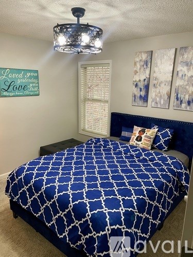 A bed with a blue and white comforter is in the foreground of a room with a chandelier and a wall hanging.