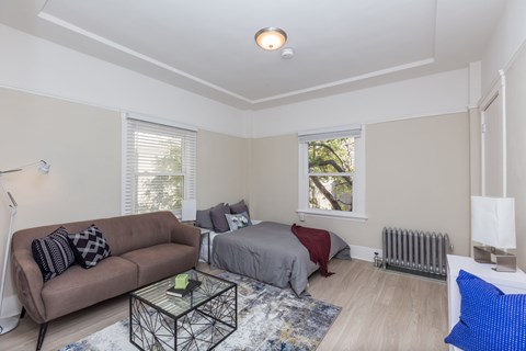 A living room with a brown couch, a glass coffee table, and a bed with a red blanket.