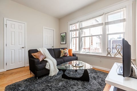 A black couch with a white blanket and pillows sits in a living room with a glass coffee table in front of it.