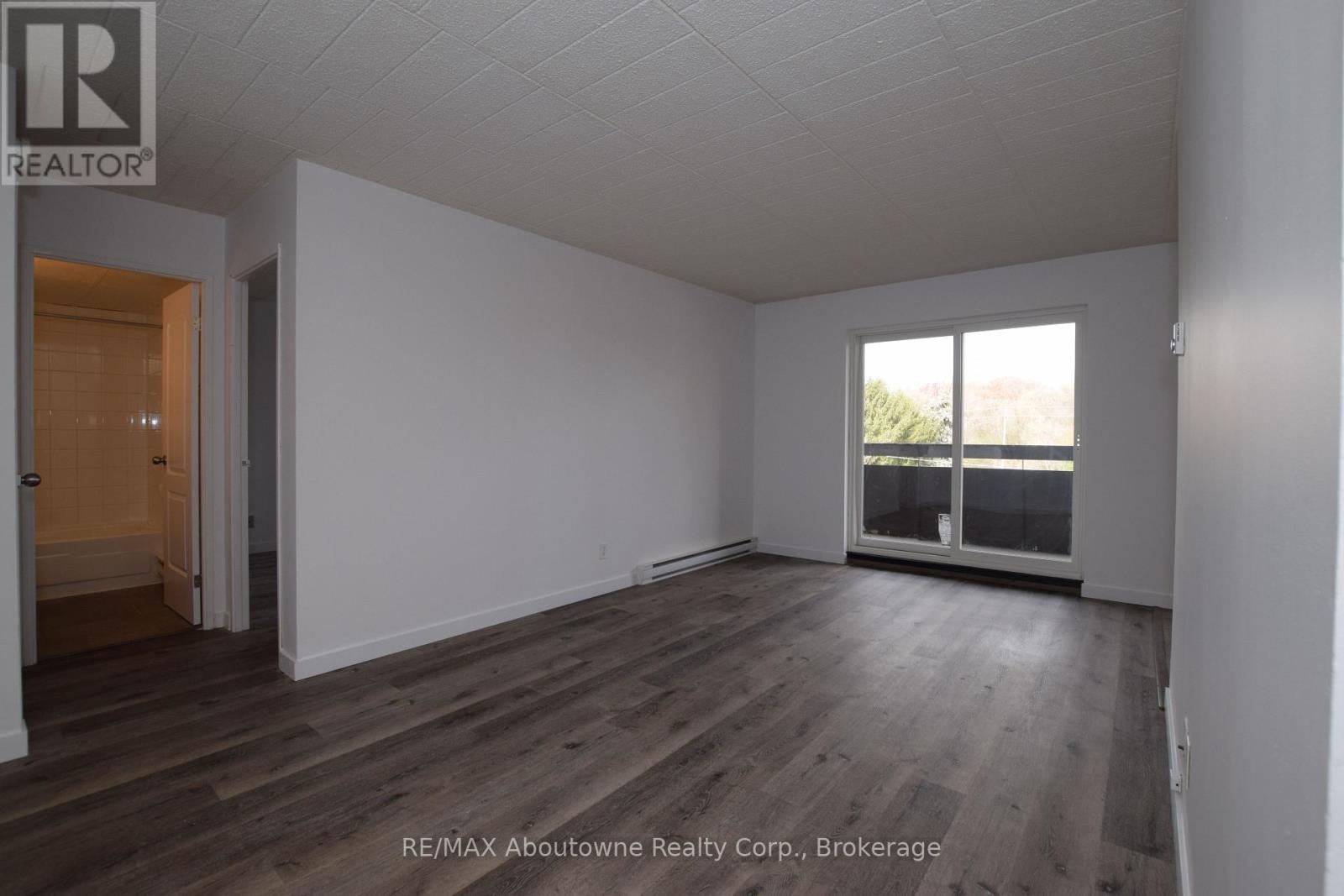 A room with wooden flooring and white walls, with a door on the left and a window on the right.