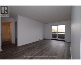 A room with wooden flooring and white walls, with a door on the left and a window on the right.