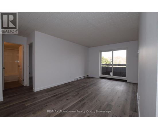 A room with wooden flooring and white walls, with a door on the left and a window on the right.