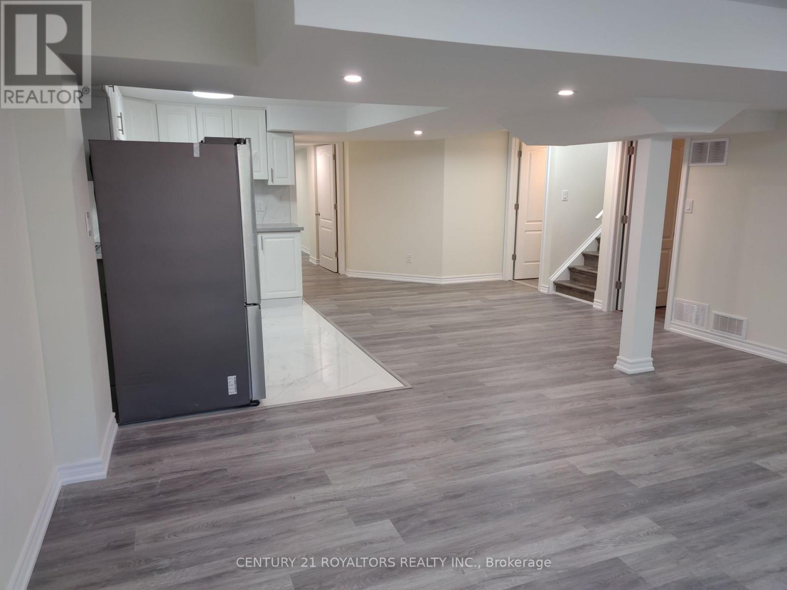 A spacious room with a grey floor and white walls, featuring a large open space with a staircase and a refrigerator.