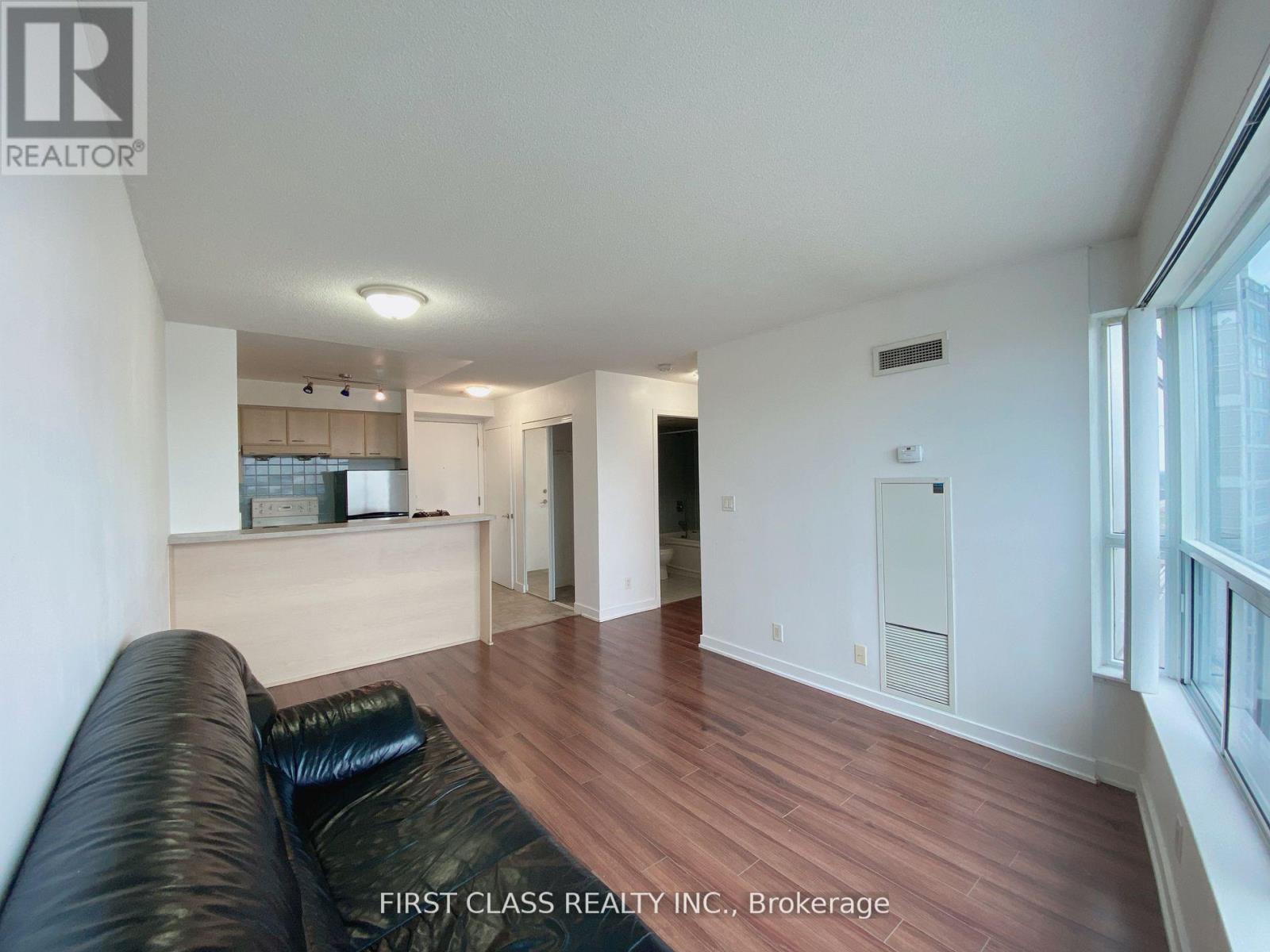 A black leather couch sits in a spacious living room with wood flooring and white walls.