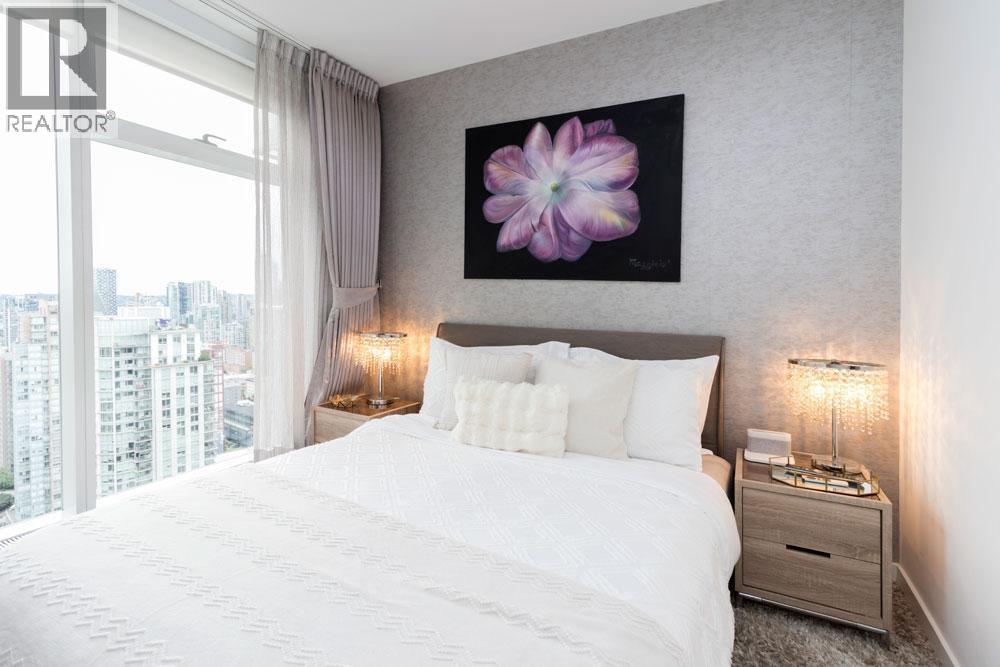 A bedroom with a large bed, a nightstand with a lamp, and a framed picture of a flower on the wall.