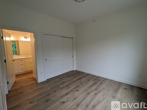 A room with wooden flooring and white walls, with a kitchen visible through a door.
