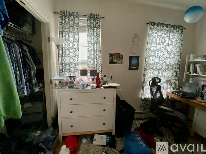 A messy bedroom with a white dresser and a window with blue curtains.