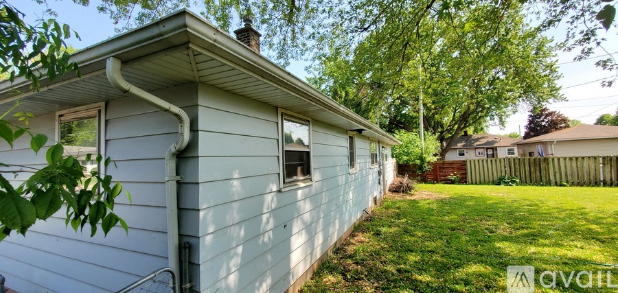 A small house with a grey siding and a white gutter is surrounded by a green lawn and trees.