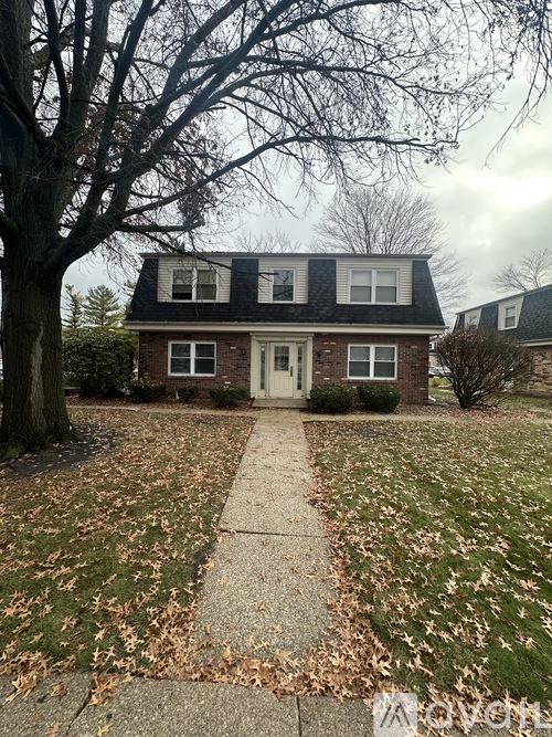 A house with a grey roof and a white door is surrounded by a lawn with fallen leaves.