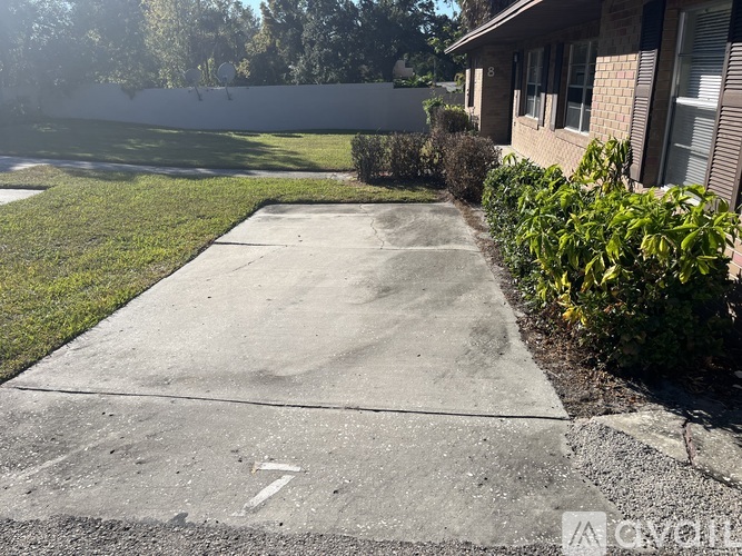 A concrete driveway leads to a house with a white wall and green bushes on the side.