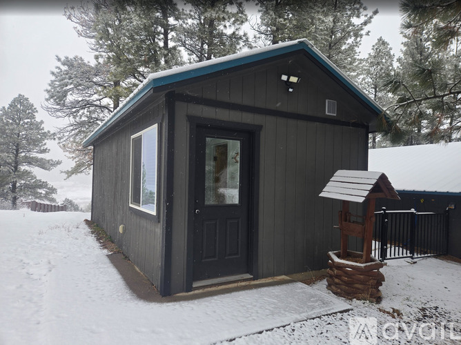 A small cabin sits in the snow with a wooden fence and trees in the background.