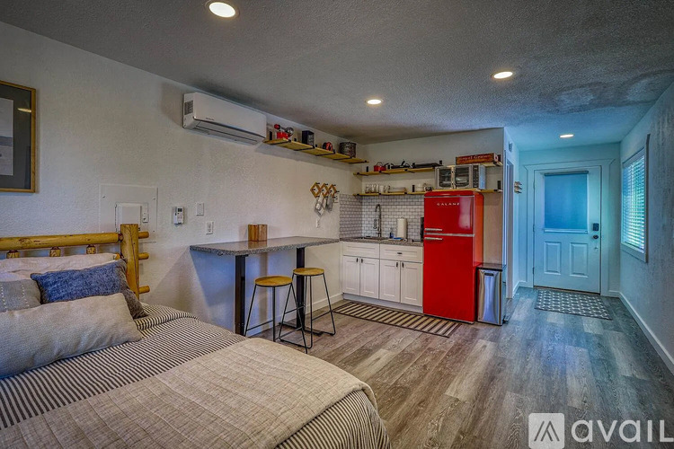 A bedroom with a bed, a table, and a refrigerator in the kitchen area.