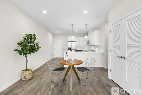 A room with a round table and chairs, a potted plant, and a kitchen area in the background.