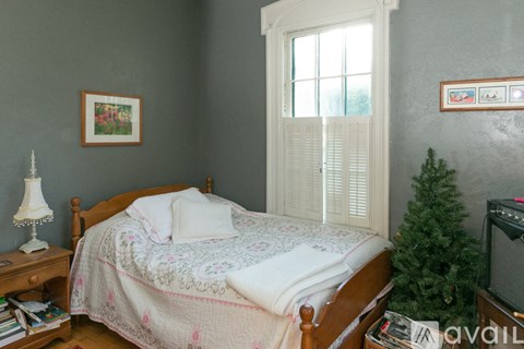 A bedroom with a bed, a nightstand with a lamp, a window with white shutters, a framed picture on the wall, and a small Christmas tree.