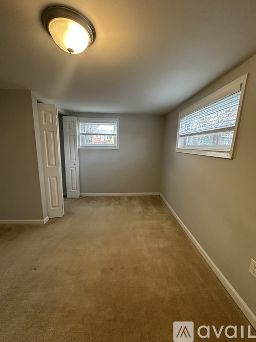 A spacious room with beige carpeting and white walls, featuring a bathroom with a toilet and sink visible through a glass door.