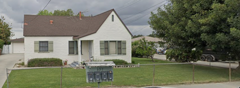 A white house with a brown roof and a grey trash can in front of it.