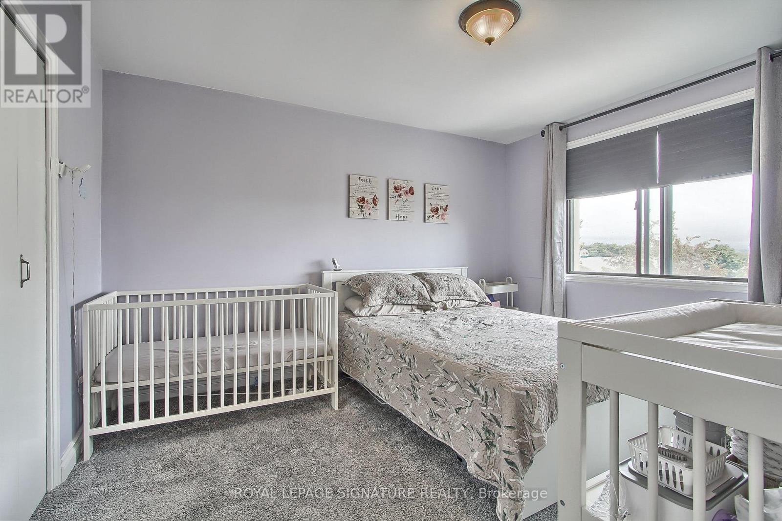 A white crib sits in a bedroom with a grey carpet and a white bed.