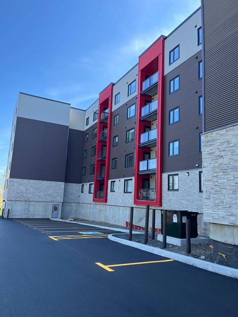 A modern building with a red and grey facade stands next to a parking lot.