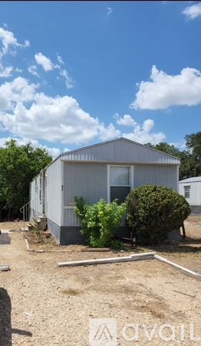 A single-story house with a grey roof and white walls is surrounded by a few bushes and trees.