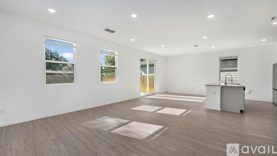 A spacious room with wooden flooring and white walls, featuring a kitchenette and a large window.