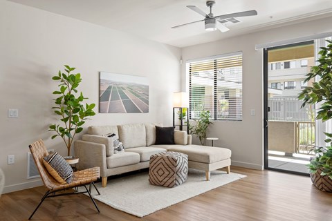 A living room with a beige couch, a chair, a small ottoman, a rug, a potted plant, and a painting on the wall.