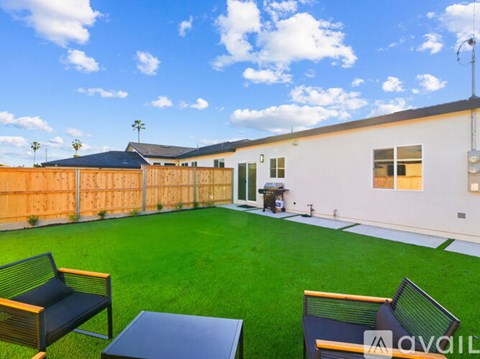 A backyard with a white building, a wooden fence, and a green lawn with two chairs and a table.