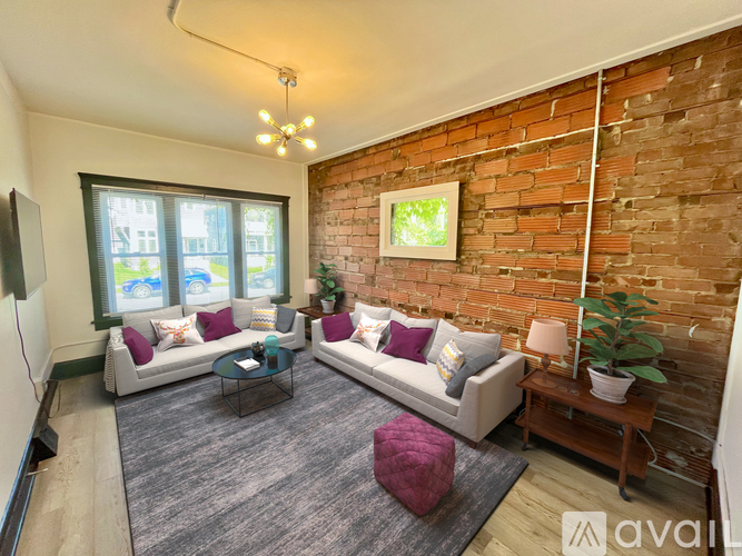 A living room with a grey couch, a red ottoman, and a brick wall.