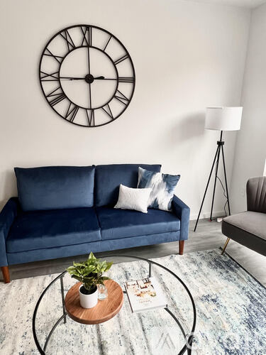 A living room with a blue couch, a round clock on the wall, and a small potted plant on a coffee table.