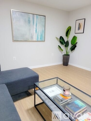 A living room with a grey sofa, a glass coffee table with books and a candle, a potted plant and a painting on the wall.