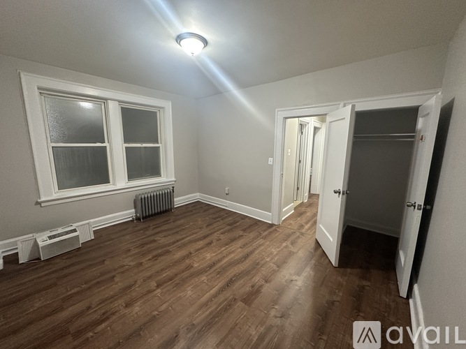 A room with wooden floors and white walls, featuring a window, a radiator, and a door.