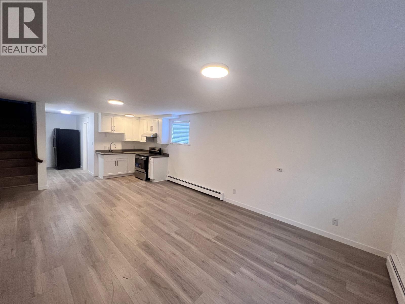 A large empty room with wooden flooring and a kitchen area in the background.