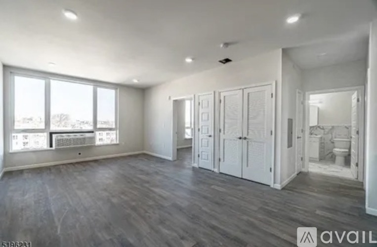 A spacious room with wooden floors and white walls, featuring a large window with a view of trees and buildings outside.