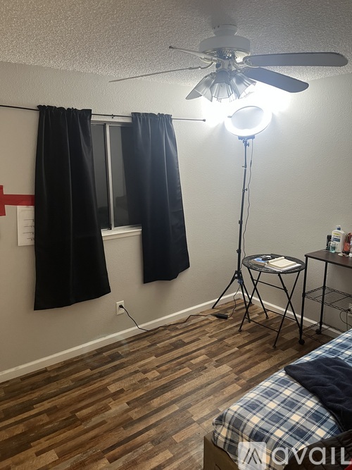 A room with a bed, a fan, a table, a chair, a window with curtains, and a lighting setup.