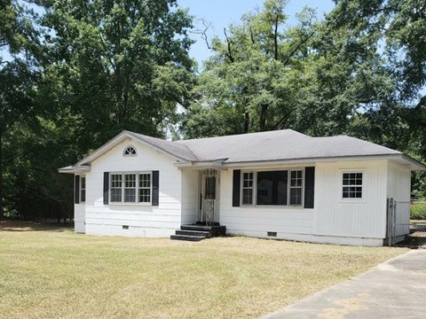 A small white house with a grey roof is surrounded by green trees.