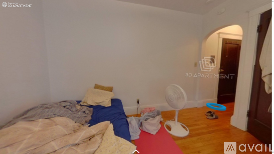 A room with a bed, a fan, and a red mat on the floor.