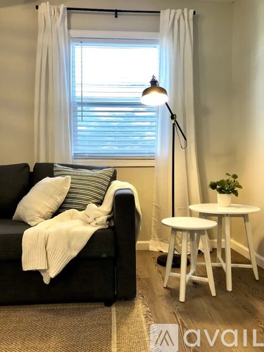 A black couch with a white blanket and pillows sits in front of a window with white curtains.