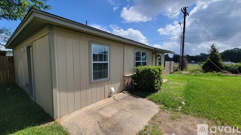 A small house with a brown siding and a white door is surrounded by a grassy area and a dirt path.