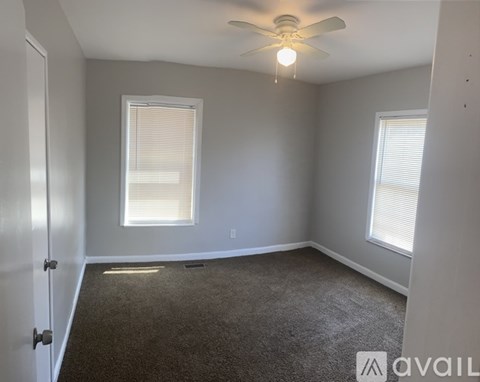 A room with a carpeted floor, a ceiling fan, and two windows with blinds.
