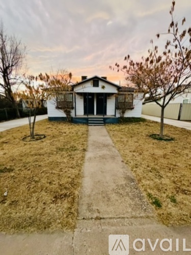 A white house with a black door and windows is surrounded by a grassy area and a concrete pathway leading to the front door.