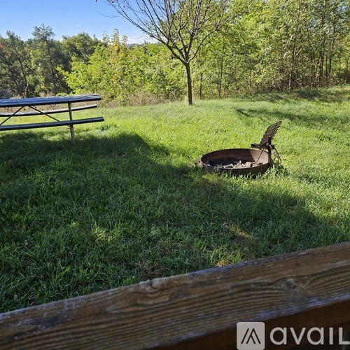 A picnic table is in the foreground of a grassy field with a fire pit and tree in the background.