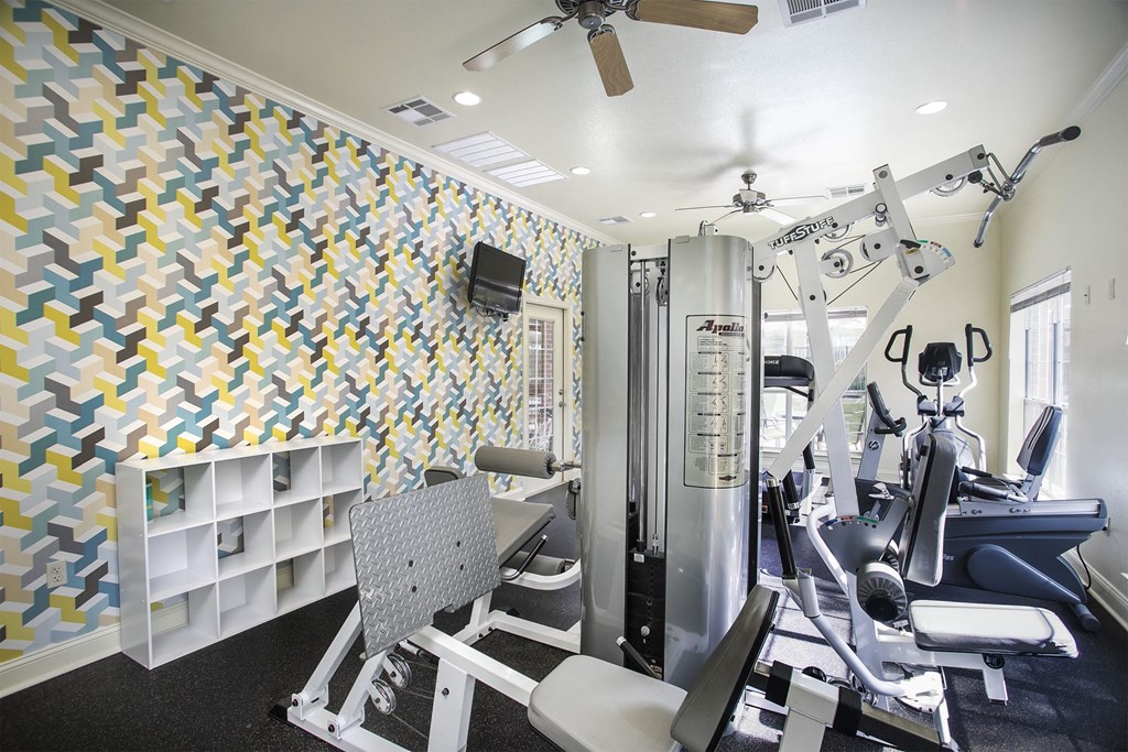Tyler, TX Apartments - Community Fitness Center with Weight Machines, a Ceiling Fan, and Large Windows.