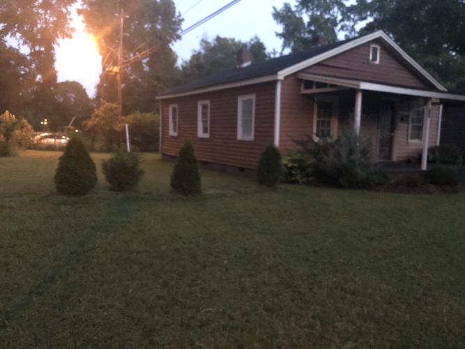 A house with a brown siding and a white trim is surrounded by a grassy lawn and small trees.