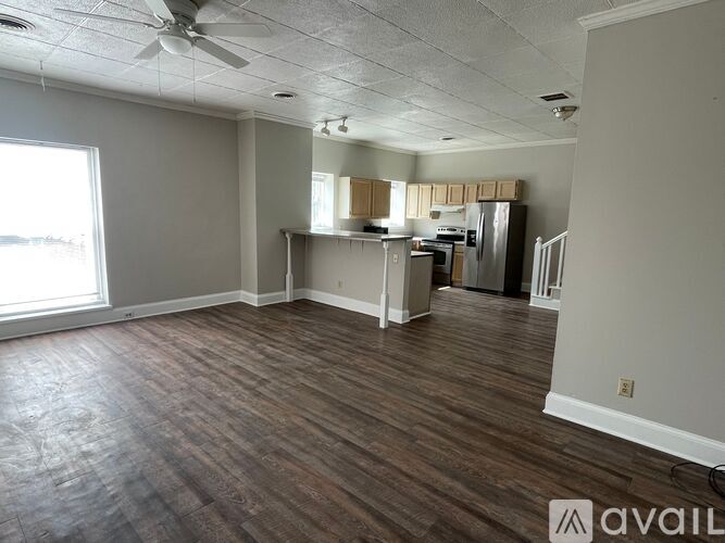 A room with a fan and a kitchen area in the background.