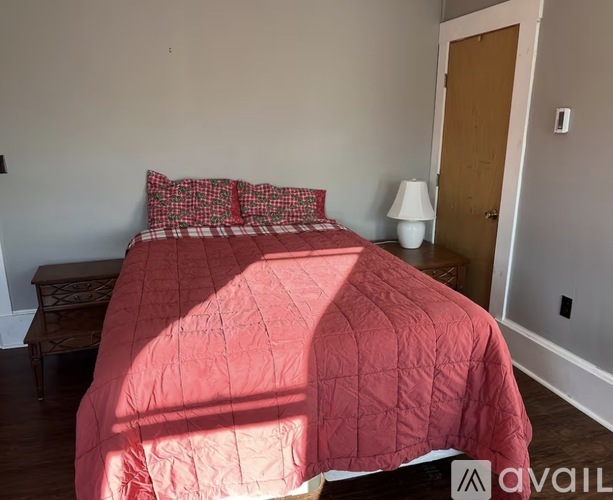 A bed with a red comforter and matching pillow cases sits in a room with a wooden floor and white walls.