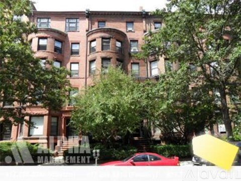A red car is parked in front of a brown building with a green tree in front of it.