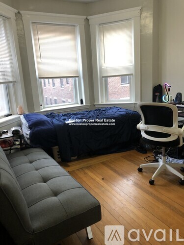A bedroom with a bed covered in a blue sheet, a grey ottoman, and a white chair.