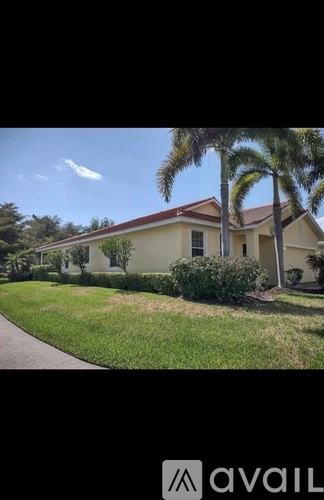 A house with a red roof and a white wall is surrounded by green grass and palm trees.